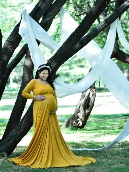 A full-length portrait of an expectant mother in a flowing yellow gown, standing by a tree draped with white fabric. This shot showcases how I use simple props to add elegance to outdoor settings.