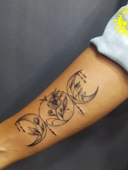 The triple moon symbol, representing the maiden, mother, and crone. This forearm piece is interwoven with flowers, adding a touch of natural beauty to the spiritual symbol.