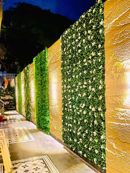 A full view of a long compound wall featuring alternating panels of stone, dense green foliage, and white flowering plants. This design breaks up the monotony of a long wall and adds significant curb appeal.