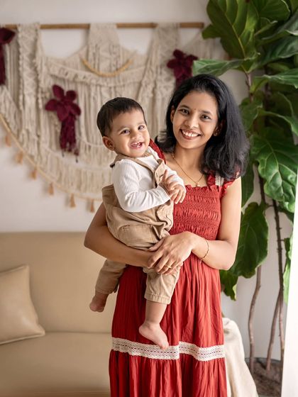 A mother holds her smiling son in a warm, inviting living room setting. The rich red of her dress contrasts beautifully with the neutral tones of the studio.
