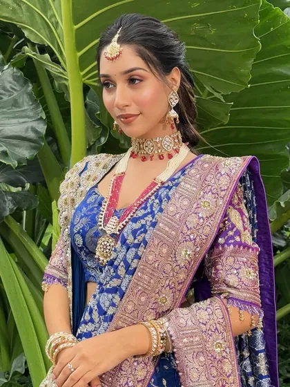 A model showcasing a royal blue and purple bridal look, perfect for a grand Sangeet or reception. The makeup features pink-toned eyeshadow and glowing skin.