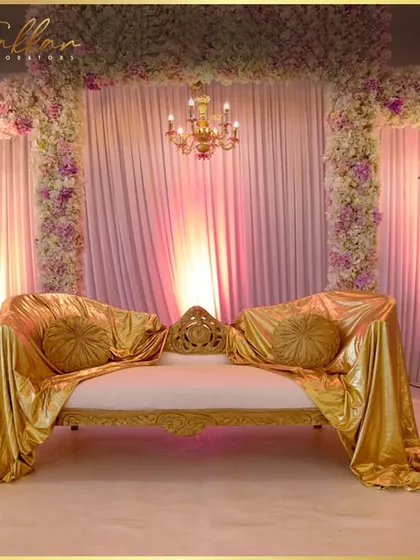 A beautiful reception stage with a gold satin-draped sofa. The backdrop of pink flowers and chandeliers adds to the elegant and romantic feel of the setup.