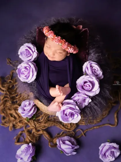 A sleeping newborn is nestled in a basket, surrounded by beautiful purple roses against a rich purple backdrop. The floral crown adds a final touch to this artistic composition.