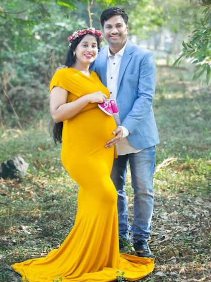 A classic maternity pose with the couple standing together in a forest setting, holding a pair of tiny pink shoes. The mother's yellow gown and floral crown create a beautiful look.