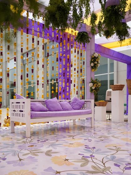 A relaxed seating area for a Mehendi or Haldi. A simple white bench with lavender cushions is set against a backdrop of hanging marigold strings and a beautiful floral-patterned floor.