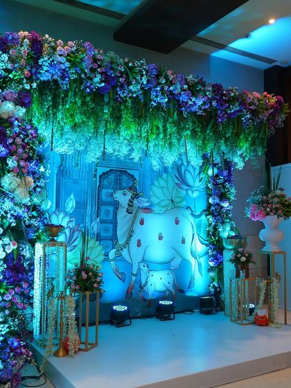A beautiful stage backdrop for a Krishna-themed baby shower. The design features a Pichwai-style painting of a sacred cow under a canopy of hanging blue and purple flowers, creating a divine and artistic focal point.