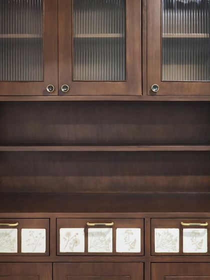 A crockery unit combining fluted glass, dark-stained wood, and custom floral tiles. This piece balances display and storage, with the handmade tiles adding a personal, artistic touch to the functional drawers.