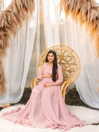 A peaceful portrait of an expectant mother in a pink gown, seated on a wicker chair. The backdrop of sheer curtains and pampas grass creates a soft and natural-looking studio environment.