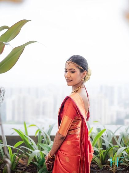 A serene portrait of a South Indian bride in a traditional red saree, looking over her shoulder with the city skyline in the background.