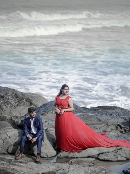 A dramatic pre-wedding shot on the rocks. The long red trail of the gown cascades over the stones, creating a stunning visual against the ocean.