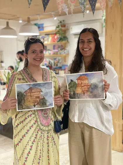 Two friends proudly hold up their watercolor landscape paintings. Our mentors guide you through the process, so you can create something you're truly happy with.