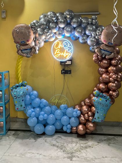 A stylish baby shower backdrop using a mix of metallic bronze and classic blue balloons. The 'Oh Baby' neon sign in the center of the circular arch adds a trendy, modern feel.