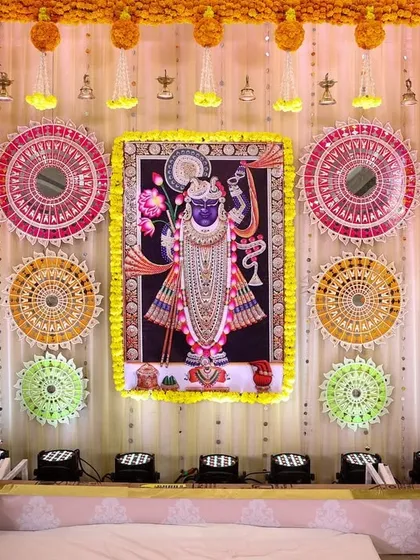 A traditional stage backdrop featuring a Shreenathji painting. The decor is enhanced with colorful thread-work panels and marigold garlands, perfect for a pre-wedding religious ceremony.