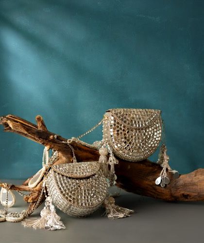 A duo of silver clutches, heavily embellished with mirror work and finished with delicate tassels. They are displayed on a piece of driftwood, highlighting their artistic quality.