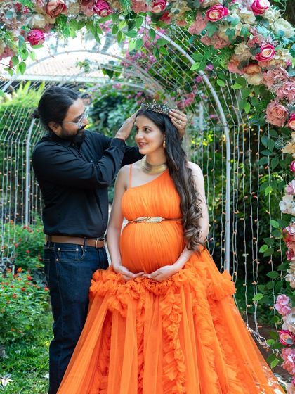 A playful and loving moment between a couple. The dad-to-be gently places a crown on his partner's head as she sits in a floral arch, both sharing a happy smile.