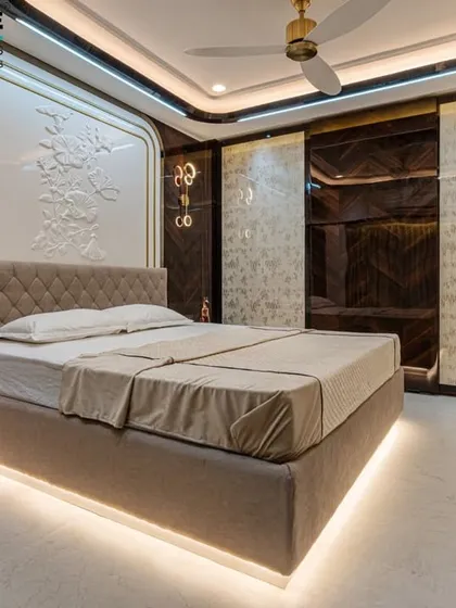 A master bedroom designed for ultimate comfort and elegance in Seawoods, Navi Mumbai. Notice the custom-designed headboard with intricate 3D relief work and gold accents, complemented by ambient lighting from the floating bed frame and designer pendant lights. The dark wood and light upholstery create a balanced, sophisticated retreat.