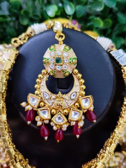 This maang tikka, part of an exclusive for-sale set, features a crescent design with white enamel work and red bead drops.