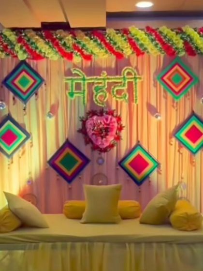 A colorful Mehandi seating area with a backdrop of light-colored drapes and vibrant geometric hangings. The low gadda seating with yellow cushions is perfect for a relaxed function.