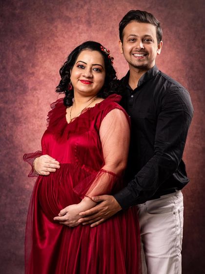 A classic studio maternity portrait against a textured backdrop. The mother-to-be is radiant in a red gown, creating a timeless and elegant image celebrating her pregnancy.
