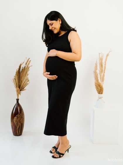 A classic and elegant full-length studio portrait. The simple black dress and minimalist background highlight the beautiful shape of her baby bump.