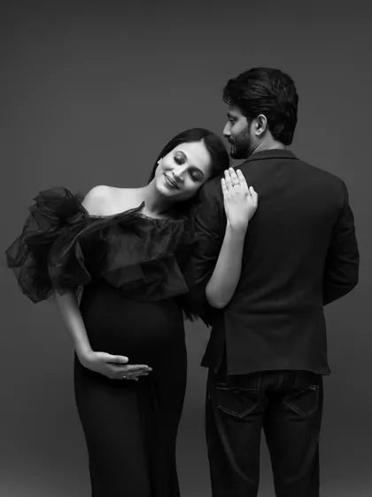 A timeless black and white maternity portrait. The expecting mother, in an elegant black gown, rests her head peacefully on her partner's shoulder, creating a classic and intimate image of their shared anticipation.