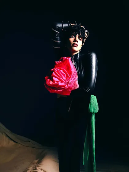 A darker, more dramatic look from 'My Flatmate's Wardrobe.' The model holds a large pink fabric flower, contrasting with the black leather and dark background.