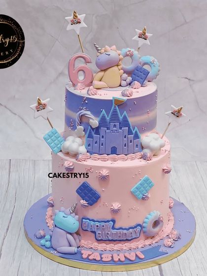 An enchanted two-tier unicorn castle cake. This black forest cake features adorable fondant unicorns, a castle, and sweet details like mini chocolate bars and donuts, all in a lovely pink and purple palette.