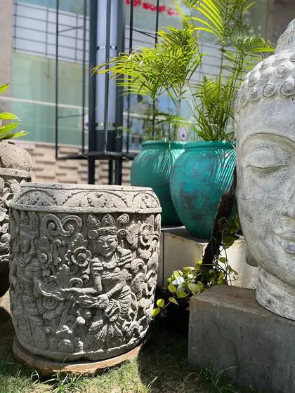 A study in texture and form. This image features an intricately carved stone planter alongside the serene, weathered face of a large Buddha head statue, with vibrant planters in the background.