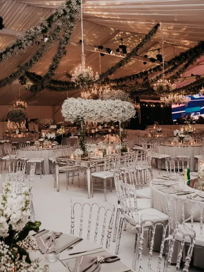 An all-white reception hall with mirrored tables, clear chairs, and lush white floral centerpieces. The ceiling is draped with greenery and chandeliers, creating a breathtakingly elegant space.