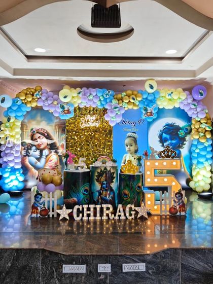 For this 5th birthday, I created an elaborate Little Krishna theme on a full stage. The decoration features two large printed backdrops, a central gold shimmer panel, a multi-colored balloon arch, character statues, and illuminated marquee letters for the child's name.