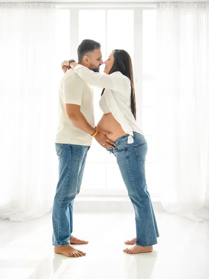 A full-length portrait of an expecting couple in matching white and denim. This high-key studio shot creates a fresh, modern, and timeless look that beautifully showcases the baby bump and the couple's bond.
