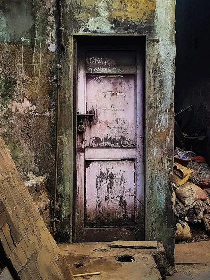 A faded pink door stands in a dark, grimy corridor, its surface covered in scratches and markings. This moody photograph captures a sense of mystery and the passage of time in an urban alley.
