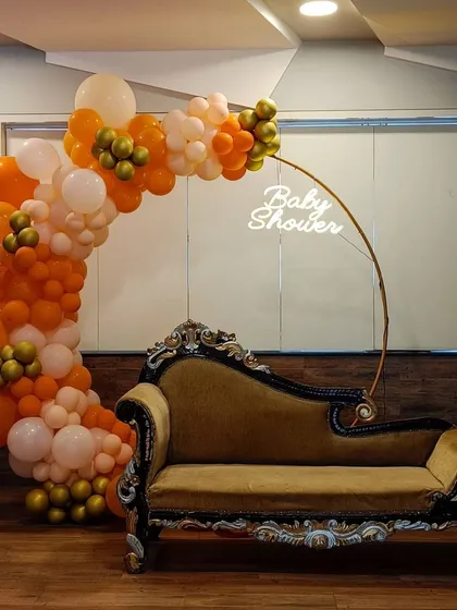 We create a special spot for the mom-to-be with this modern baby shower setup. It features a lush organic balloon arch in orange, white, and gold, a custom "Baby Shower" neon sign, and an elegant chaise lounge for comfort and perfect photos.