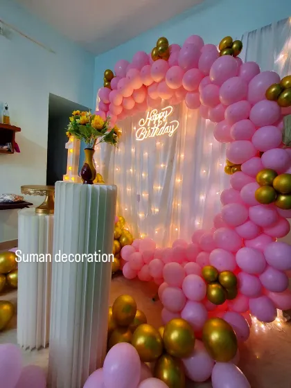 An intimate at-home birthday decoration. This setup includes a semi-arch of pink and gold balloons against a white curtain with fairy lights, creating a warm and festive corner.