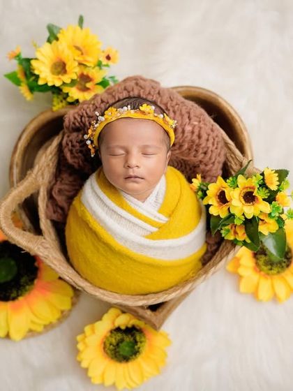 A sweet newborn wrapped in yellow and white, peacefully sleeping in a basket surrounded by bright yellow sunflowers on a fluffy white rug.