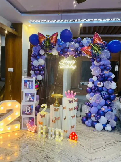 A beautiful butterfly-themed second birthday at home. This setup features a purple and blue balloon arch with large butterfly foil balloons, a 'Happy Birthday' neon sign, and light-up marquee numbers.
