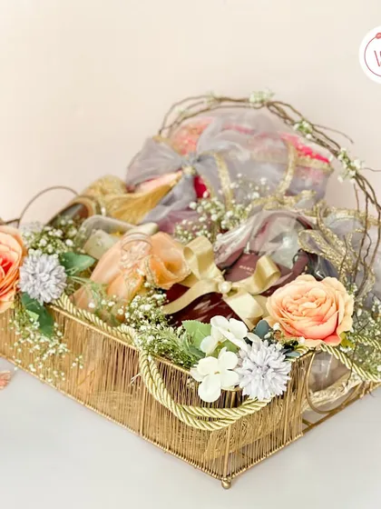 A beautiful hamper in a golden wire basket, filled with elegantly wrapped sweets and dry fruits, and adorned with peach and blue florals.