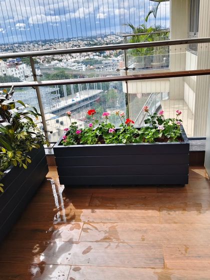Two custom rectangular planters on a wooden deck balcony. One is filled with flowering geraniums, adding a splash of color, while the other has a darker foliage plant for contrast.