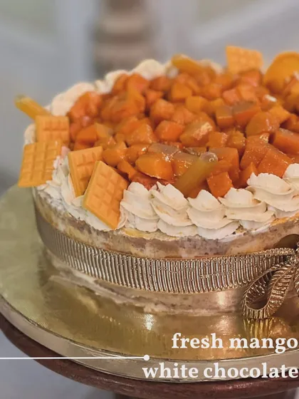 A stunning Fresh Mango & White Chocolate Cake. The cake is generously topped with fresh mango chunks and decorated with a beautiful ribbon, making it a perfect celebration cake.