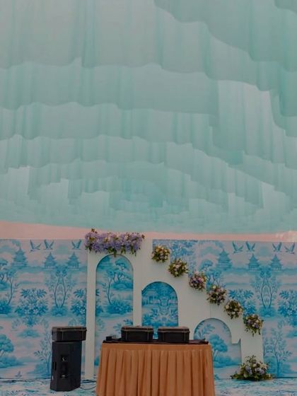 The DJ console and stage for the Dior-themed Mehendi, featuring a backdrop with the iconic blue and white print and a cascading floral arrangement, ensuring every element fits the couture theme.
