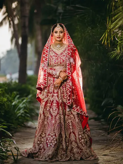 A classic, full-length portrait of the bride in her stunning red lehenga, set against a beautiful garden path. This image showcases the grandeur of her bridal attire.