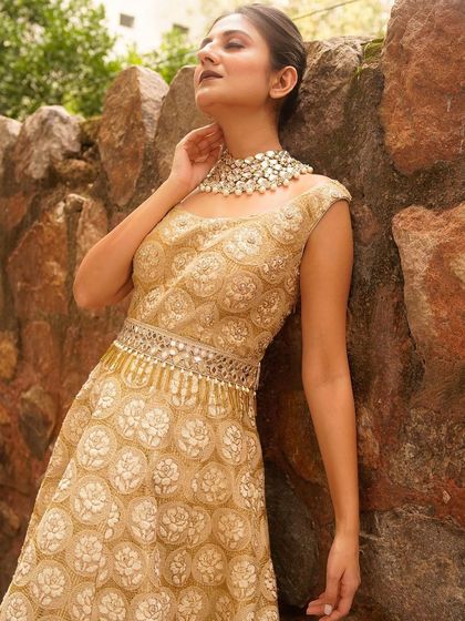 An ethereal portrait featuring a golden gown against a rustic stone wall. The sunlight filtering through the leaves adds a magical quality, highlighting the intricate texture of the dress.