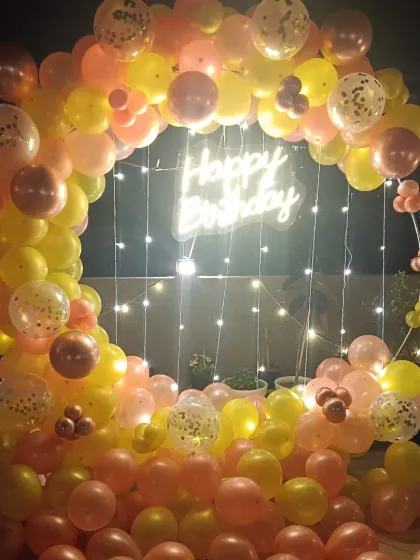 A beautiful balloon ring decoration for a terrace party at night. The yellow and pink balloons are interwoven with fairy lights, and a central "Happy Birthday" neon sign creates a warm, inviting glow.