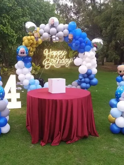 An outdoor Boss Baby themed party setup for a 4th birthday. This decoration includes a balloon arch in blue, white, and gold, character-themed balloon pillars, and a central table, perfect for a garden or backyard celebration.