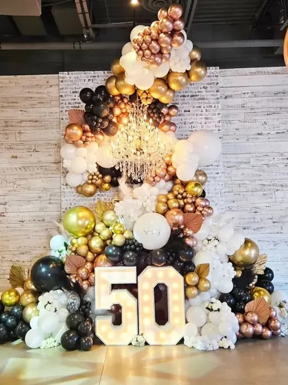 A grand and glamorous 50th birthday setup. This stunning display features a massive organic balloon arrangement in black, white, and metallics, a crystal chandelier, and a large light-up '50'.