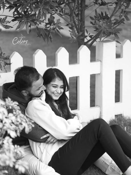 A joyful, candid black and white photo of the couple cuddling by a white picket fence.