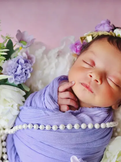 A close-up portrait of a newborn with a floral headband, peacefully asleep. The details of the pearl necklace and purple wrap add a touch of elegance.