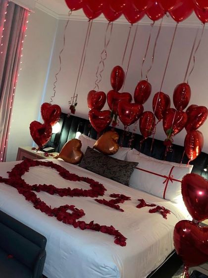 An intimate setup with "I <3 U" spelled out in rose petals on the bed, surrounded by red heart-shaped foil balloons.