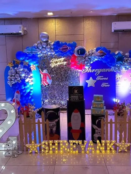 An out-of-this-world Space and Astronaut theme for a second birthday. This setup includes a silver shimmer wall, a crescent moon prop, astronaut cutouts, and a blue and silver balloon arch to create a cosmic adventure.