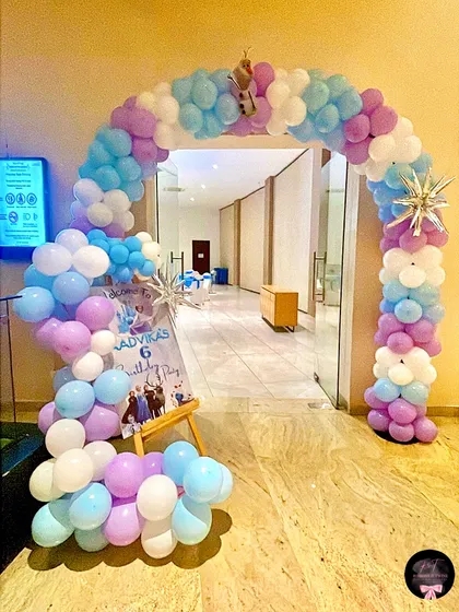 A grand balloon arch in purple, blue, and white welcomes guests to the 'Frozen' celebration. A personalized welcome sign featuring the birthday girl's name adds a special touch right at the entrance.